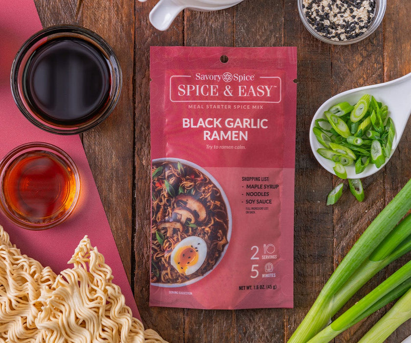 Black Garlic Ramen Spice & Easy package, top down with green onions, soy sauce, scallions, and ramen noodles arranged around it on a wood table.