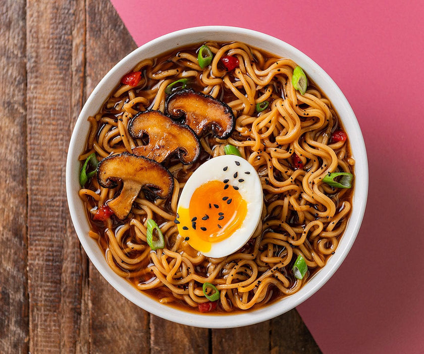 overhead shot of a bowl or ramen topped with sesame seeds, mushrooms, scallions, and a soft boiled egg. It sits on a wood table and a burgundy mat.