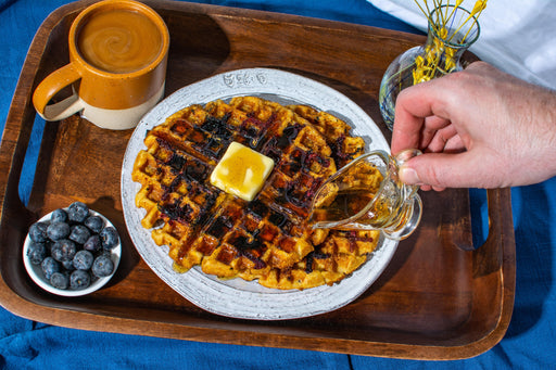 A wooden tray with blueberry lavender waffles, topped with butter and syrup, served with a mug of coffee and a bowl of fresh blueberries. 