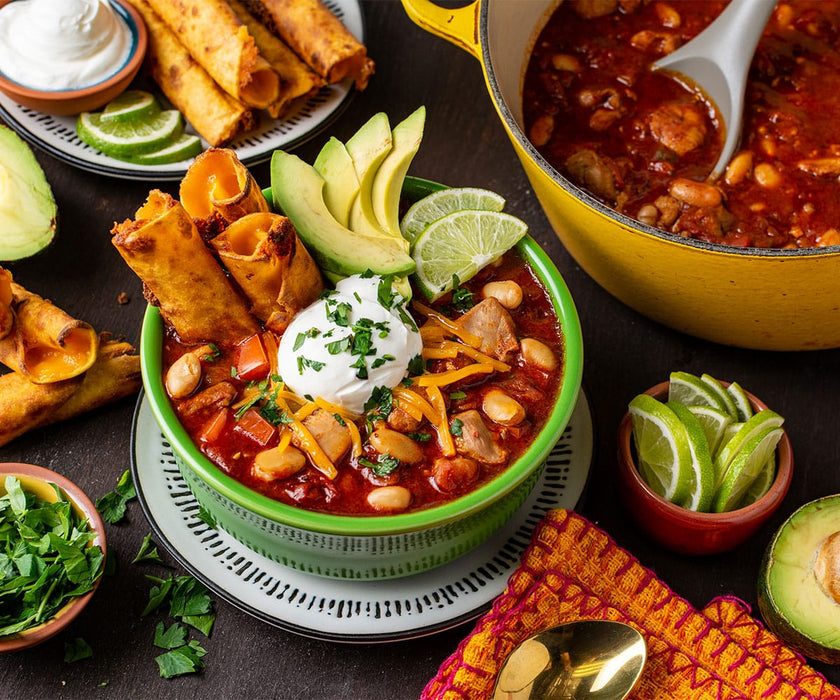 Set table with a bowl of chicken enchilada chili garnished with sour cream, avocado, cheese, lime, and tortillas, on a table with more chili, avocado, tortillas and limes