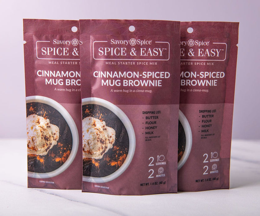 Three standing packages of the Cinnamon Spiced Mug Brownie Spice & Easy packaging on a white marble surface and a light purple background.