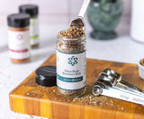 Jar of Pikes Peak Butcher's Rub on a wood cutting board with a silver measuring spoon taking spices out of the jar. 