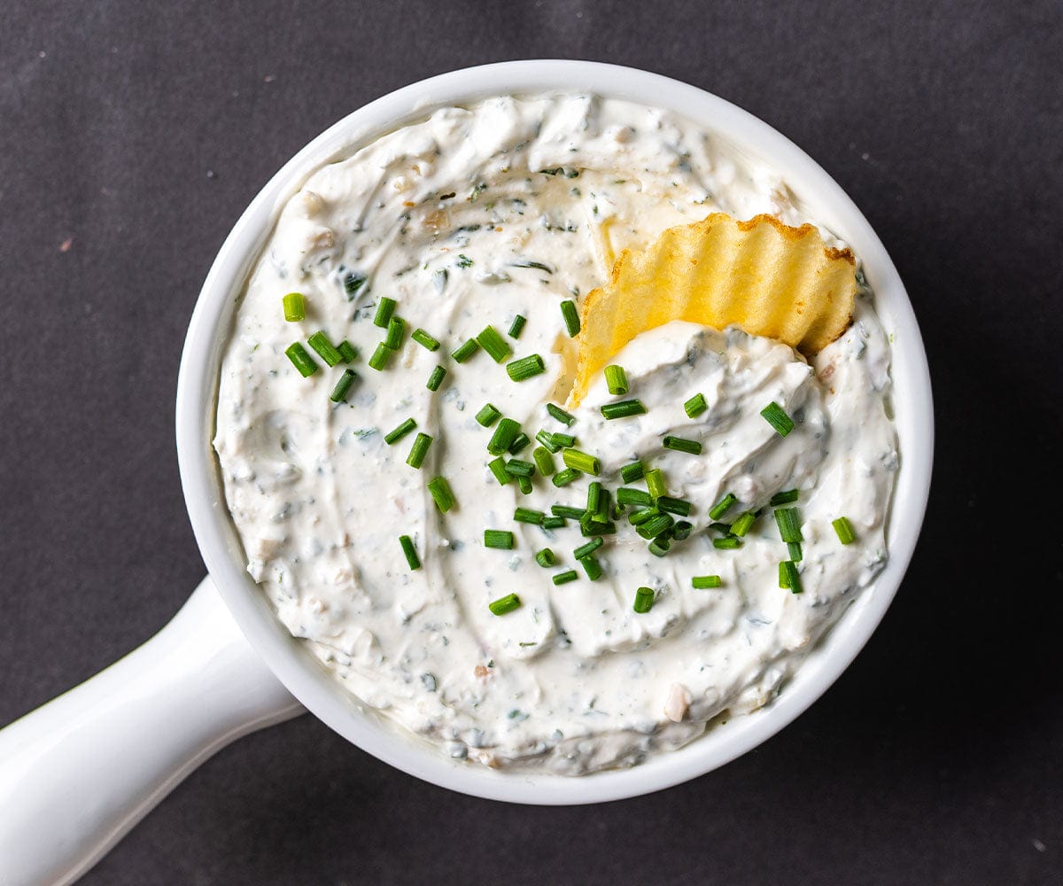 Overhead shot of a bowl of Seven Onion Dip with a chip, garnished with chives on a black background