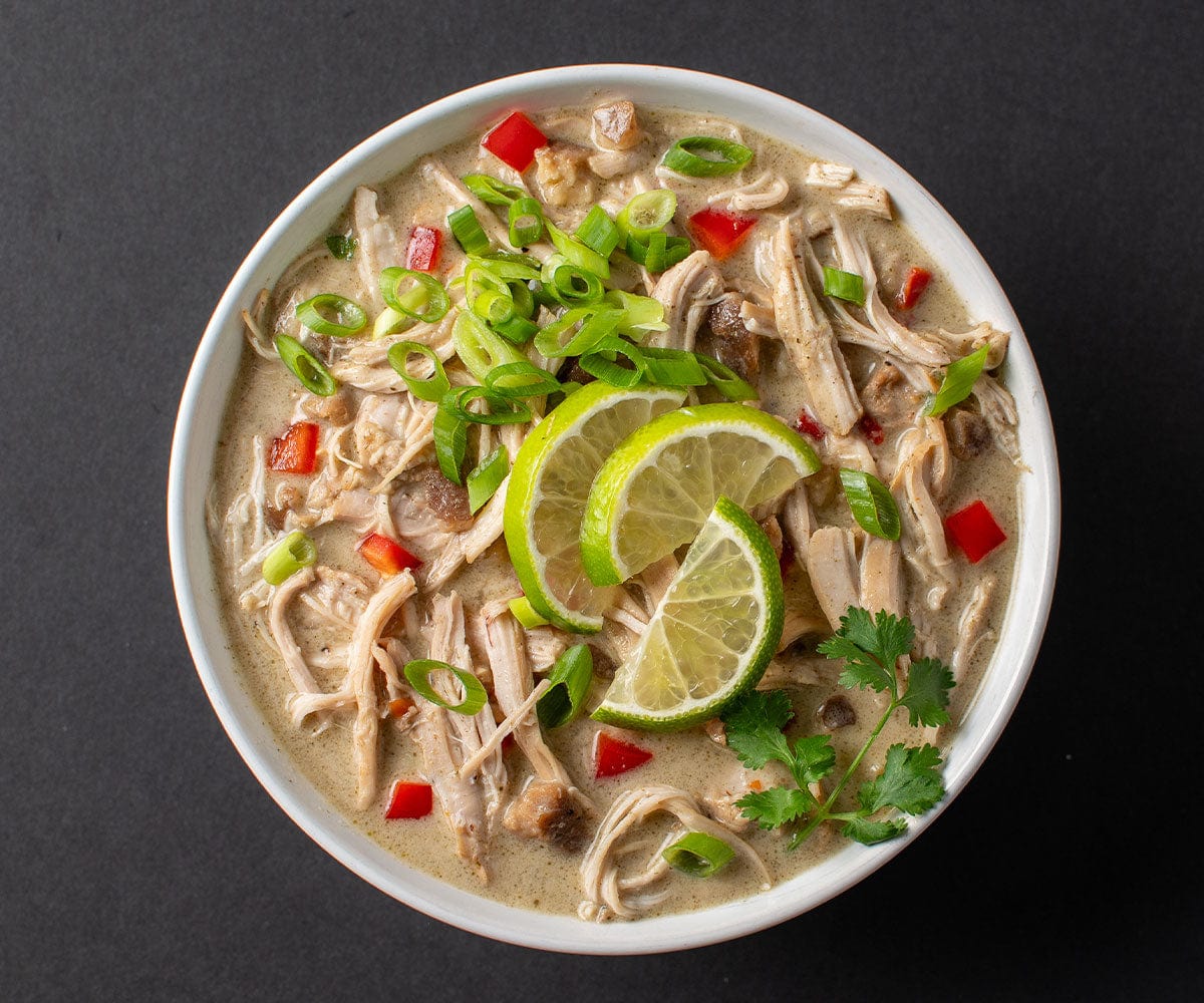 Overhead shot of a bowl of Thai Coconut Curry Soup with chicken and garnished with limes and green onions, on a black background