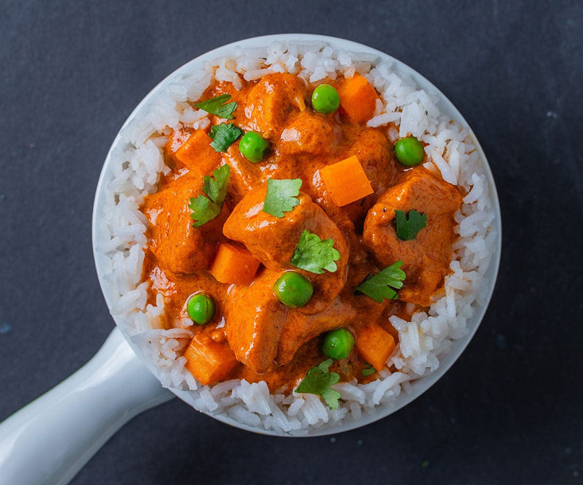 Overhead of a white bowl of Tikka Masala over rice served with peas and carrots on a black background