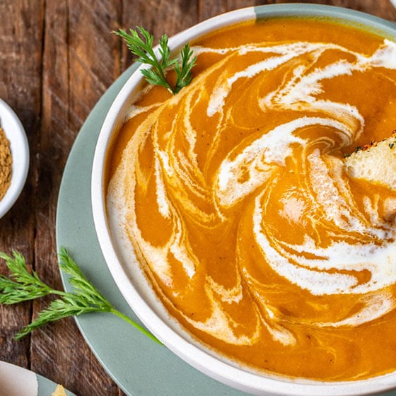 A bowl of Cumin Carrot Soup with smaller bowl of spice on the left. A hand is dipping bread into the soup.