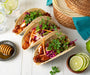 Three finished Honey Sriracha Lime Fish tacos on a table with limes and a basket of warm tortillas
