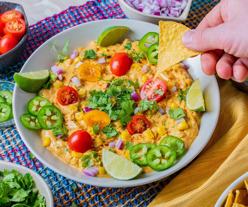 Bowl of Mexican Street Corn Dip garnished with tomatoes, jalapeno slices, and limes while someone dips a chip into it