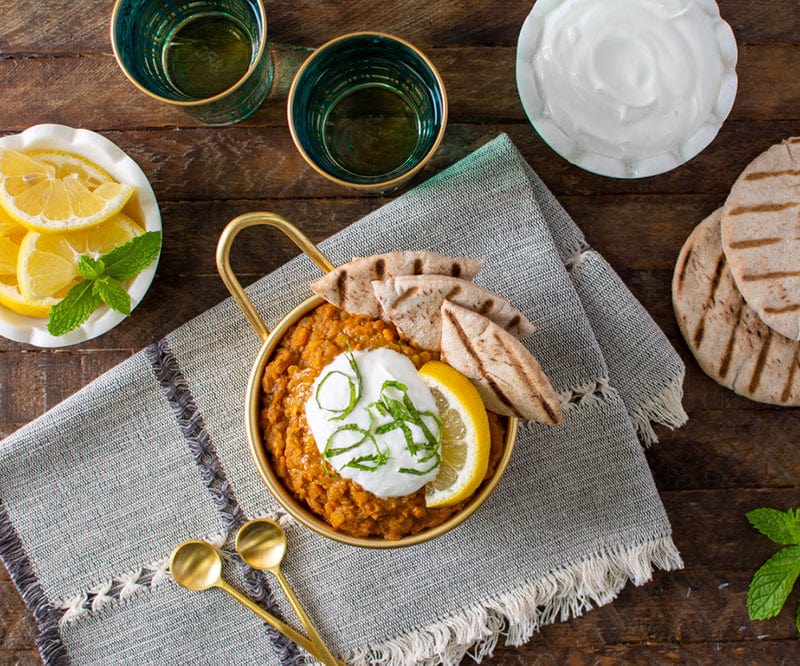 Overhead of a table set with a gold bowl of Moroccan Lentils garnished with flatbread, sour cream, lemon and herbs