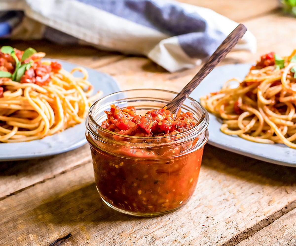 small mason jar with marinara sauce and a wooden spoon in it, in front of two plated of spaghetti 